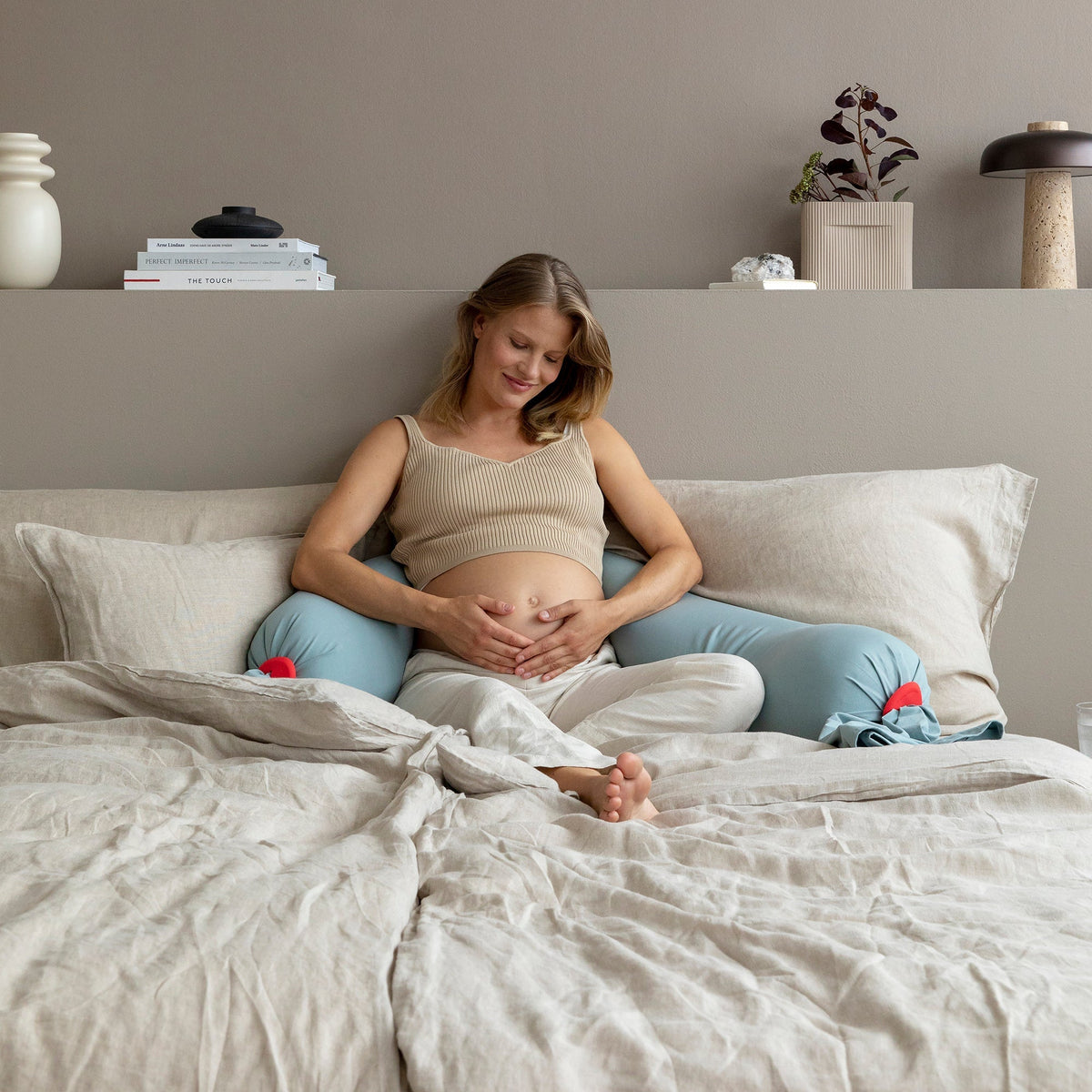 A pregnant woman sits on a bed in a neutral bedroom setting, wearing a beige tank top, light blue leggings, and white socks. The room features a warm taupe wall, a shelf with decorative items, and light beige bedding.