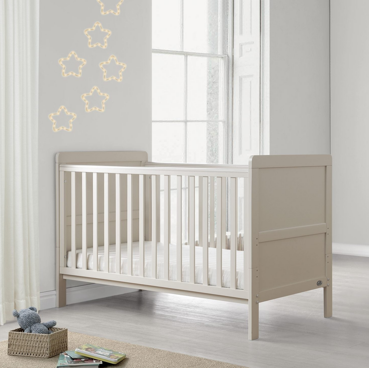 A light beige wooden baby crib in a bright, modern nursery setting with a white window, star-shaped string lights, a small wooden stool with folded blankets, and a framed alphabet print on the wall.
