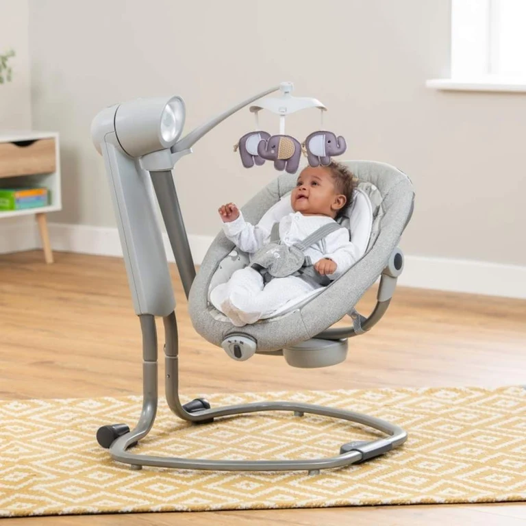 A baby in a white outfit sits in a gray electric baby swing with a mobile hanging above, placed on a patterned rug in a bright room.