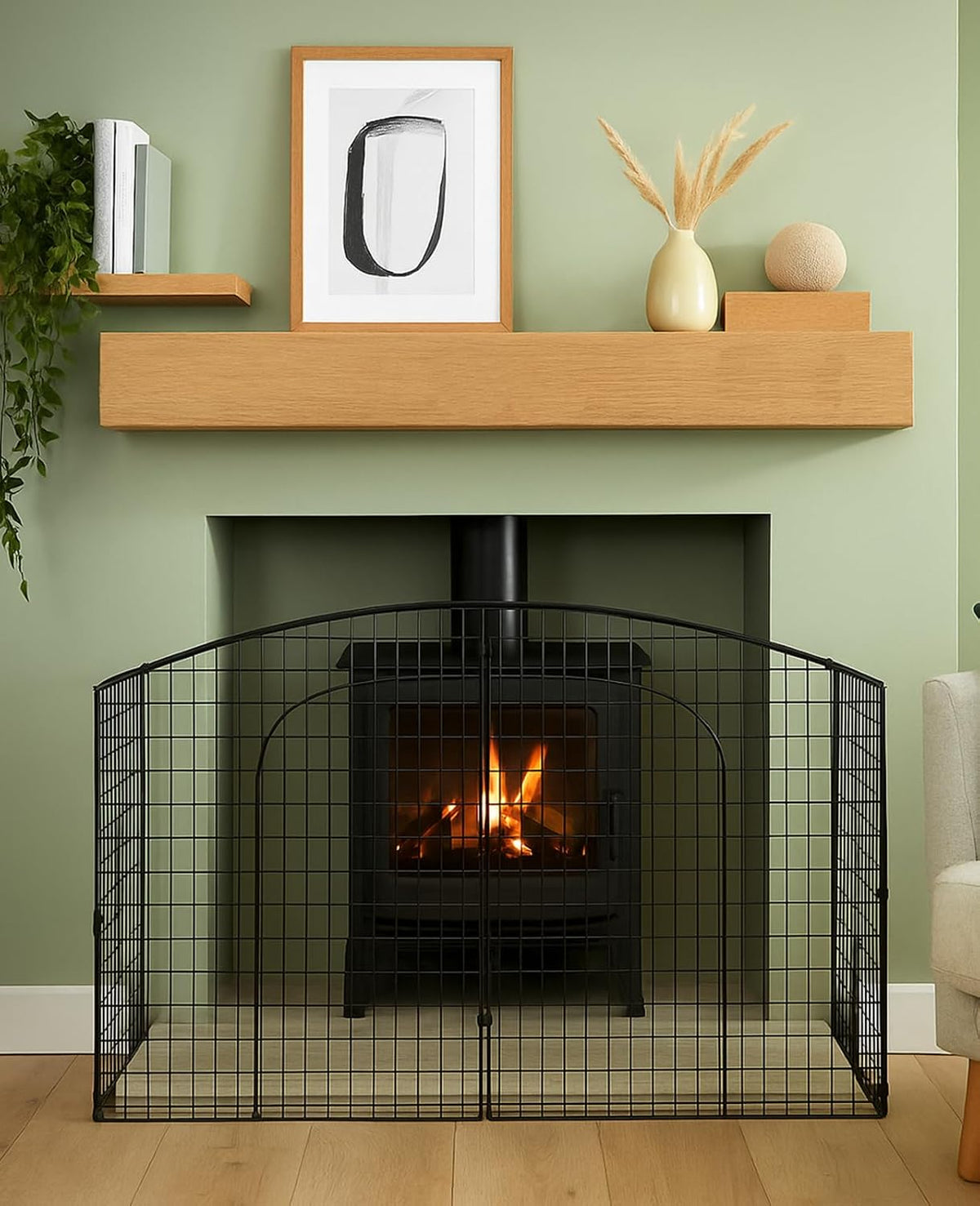 A black metal fireplace guard screen with an arched top and wire grid front, positioned in front of a black wood-burning stove inside a fireplace. The scene is set in a styled room with a sage green wall, a light wood mantel with decorative items, and a wood floor.