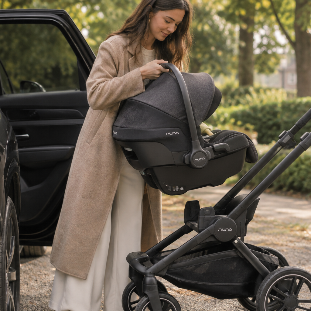 A woman in a beige coat stands beside an open car door holding a Nuna infant car seat next to a black Nuna stroller in an outdoor garden setting.