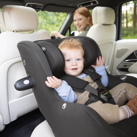 Child in a car seat with a woman driving, surrounded by greenery outside the car.