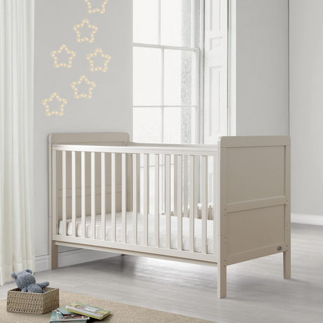 A light beige wooden baby crib in a bright, modern nursery setting with a white window, star-shaped string lights, a small wooden stool with folded blankets, and a framed alphabet print on the wall.