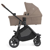 A stroller bassinet combo in taupe fabric with a black frame and wheels, shown from a side profile on a white background.