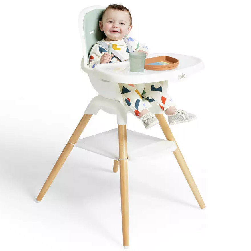 A baby seated in a white high chair with light wood legs, wearing a colorful patterned outfit. The baby is holding a small cup and there is a tray on the chair, photographed against a clean white background.