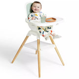 A baby seated in a white high chair with light wood legs, wearing a colorful patterned outfit. The baby is holding a small cup and there is a tray on the chair, photographed against a clean white background.