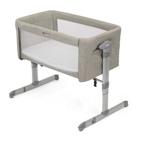 A Jola baby bassinet with a light beige textured fabric frame, white mesh sides, and a white mattress, supported by a gray adjustable metal stand on a white background.