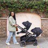 Woman pushing a black stroller with two baby seats against a brick wall.