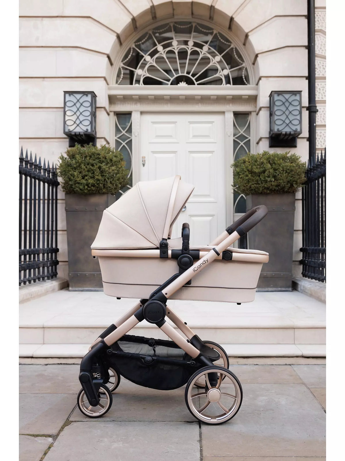 A stroller with a cream-colored bassinet and canopy, mounted on a champagne-gold and black stroller frame, parked on a stone sidewalk in front of a classic white door and stone facade.