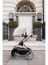 A stroller with a cream-colored bassinet and canopy, mounted on a champagne-gold and black stroller frame, parked on a stone sidewalk in front of a classic white door and stone facade.