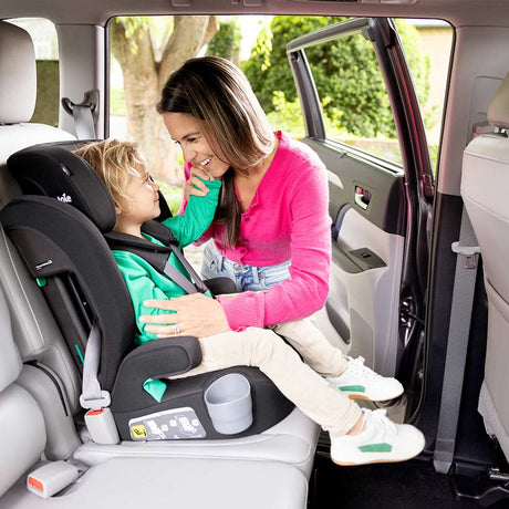 A woman in a pink sweater sits in the back of a vehicle next to a child seated in a black and gray car seat, demonstrating the safety harness and seat belt system.