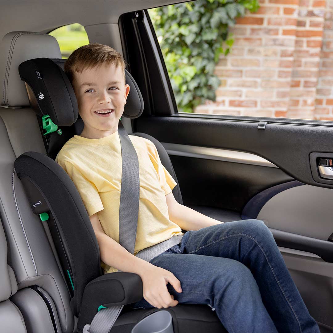A young boy wearing a yellow t-shirt and blue jeans sits secured in a black and gray child car seat installed on a car's front seat, with a window showing a brick wall and greenery outside.