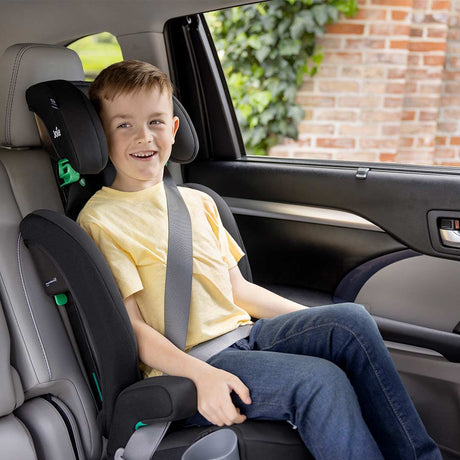 A young boy wearing a yellow t-shirt and blue jeans sits secured in a black and gray child car seat installed on a car's front seat, with a window showing a brick wall and greenery outside.