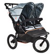 A double stroller travel system with two seats, dual canopies, and a large rear footrest, shown on a plain white background. The stroller features a black frame and wheels with gray and black fabric seats and canopies, accented by thin orange stripes.