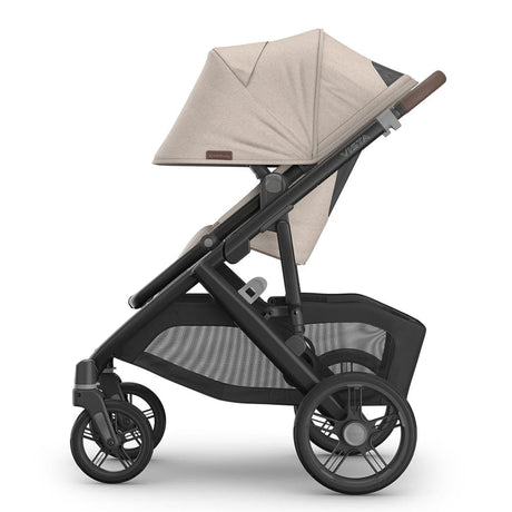 A side profile view of a stroller with a light beige fabric canopy and seat liner, a dark gray frame, and black wheels, set against a plain white background.
