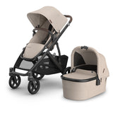 A stroller and bassinet travel system in a coordinated beige and dark gray/black colorway, shown on a clean white studio background. The stroller has a large beige canopy and seat cover with dark gray frame and black wheels, and the matching bassinet sits beside it.